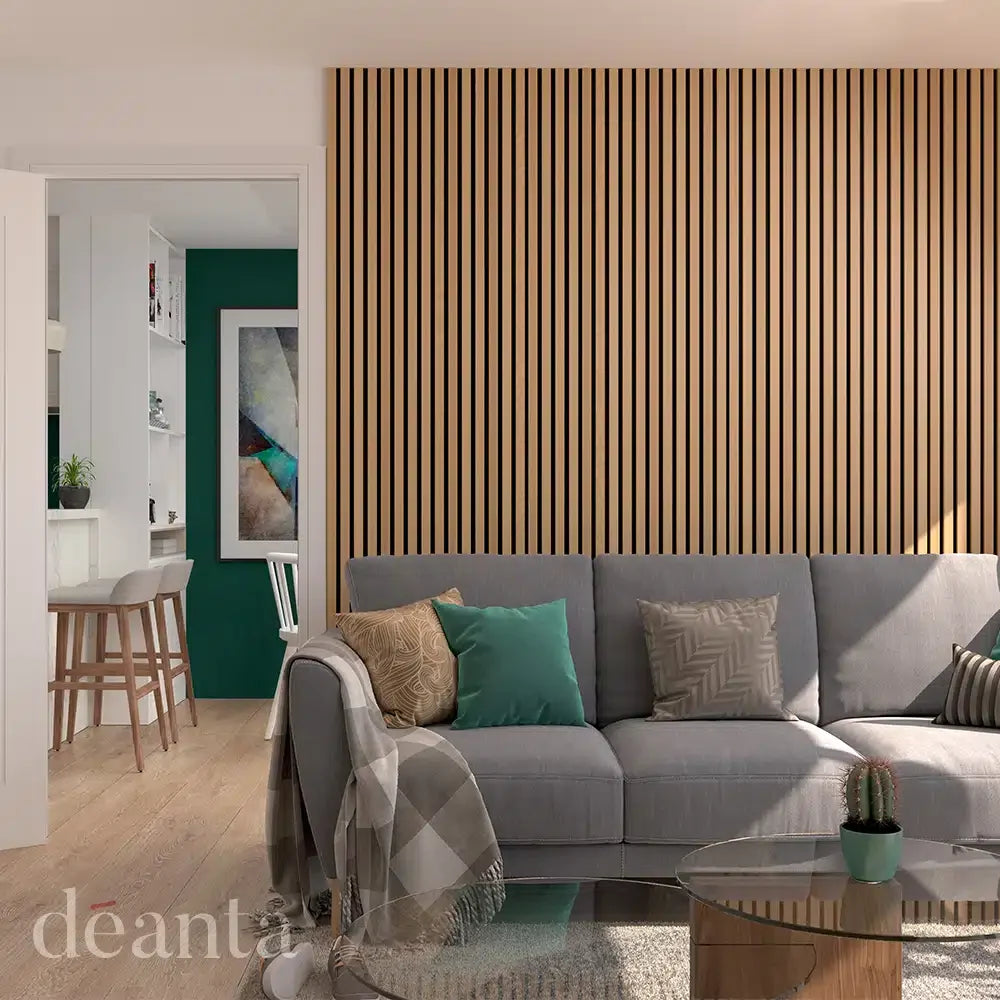 Modern living room with a gray sofa, patterned pillows, and a glass coffee table. The wall features vertical wood slats. A glimpse of a kitchen with bar stools is visible through an open door.
