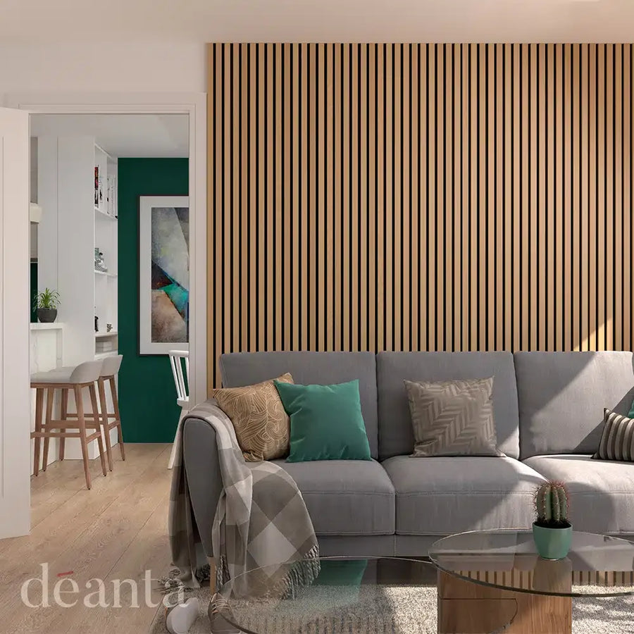 Modern living room with a gray sofa, patterned pillows, and a glass coffee table. The wall features vertical wood slats. A glimpse of a kitchen with bar stools is visible through an open door.